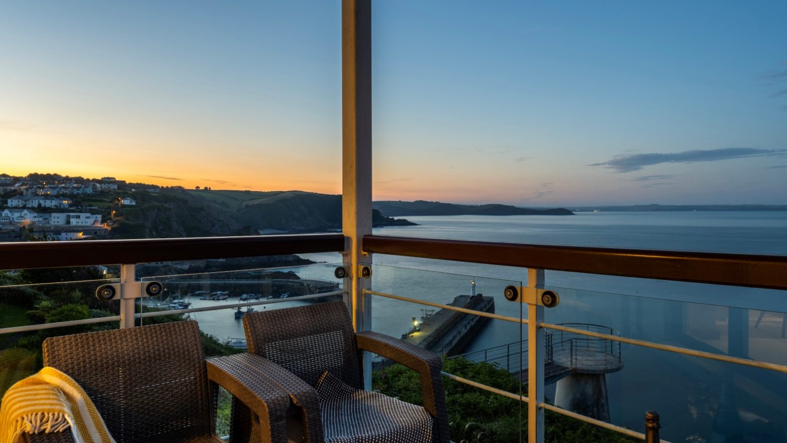 A serene view of the ocean at sunset from the Marine Point balcony. Two wicker chairs and a blanket are in the foreground, with a scenic coastline and calm waters extending into the horizon. The sky is painted in soft hues of orange and blue.