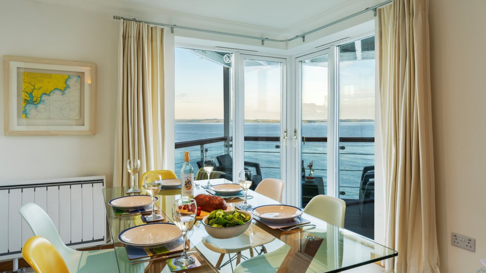 A dining table set with plates, glasses, and a meal overlooks the marine point from a balcony with an ocean view. The room is bright, featuring large windows and light curtains. A framed map adorns the wall, and yellow chairs complement the airy decor perfectly.