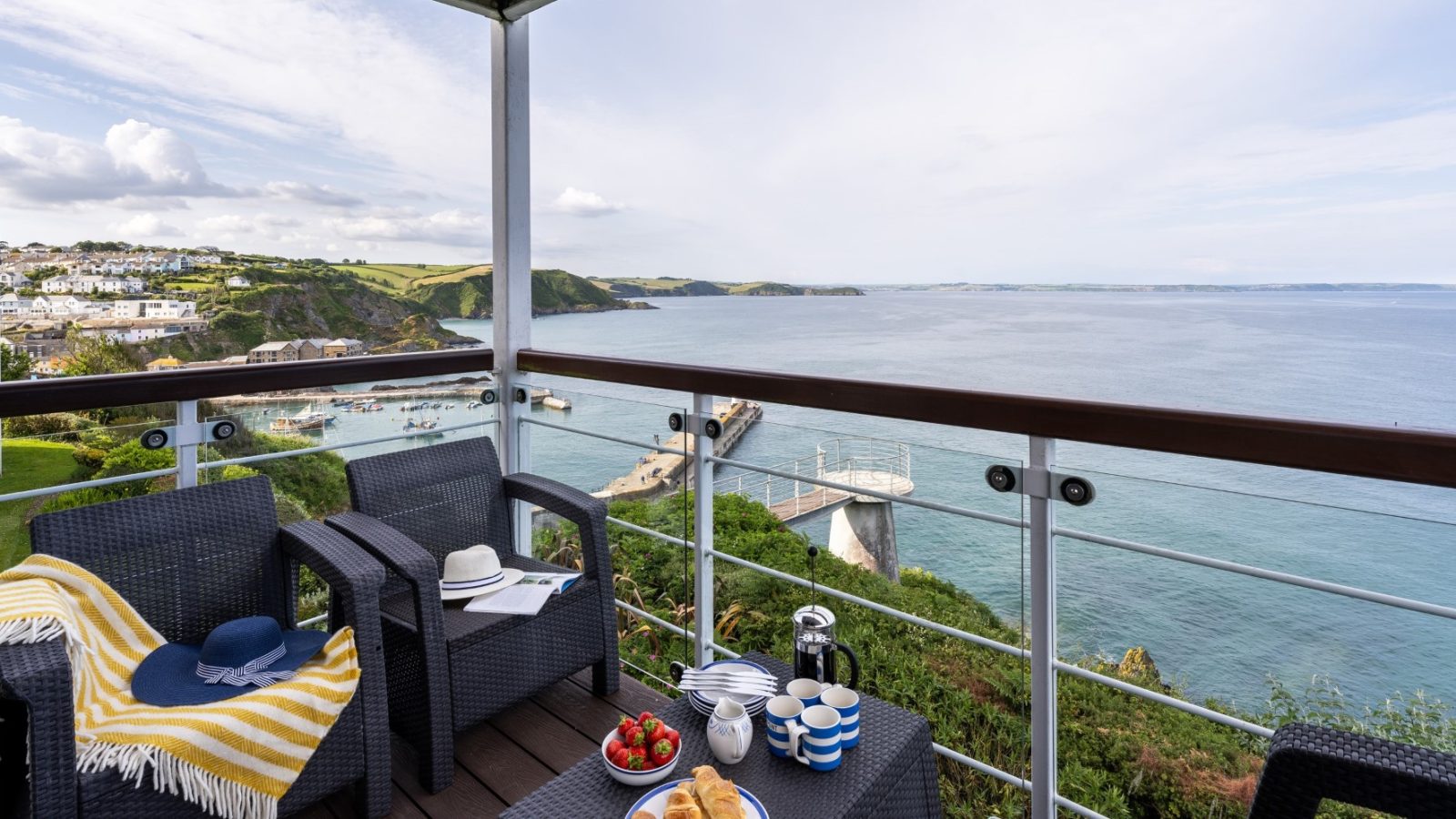 A balcony at Marine Point features two black wicker chairs and a small table set with breakfast items. A yellow striped blanket and a blue hat rest on one chair. The view overlooks a scenic coastal landscape with cliffs, greenery, and the sea under a partly cloudy sky.