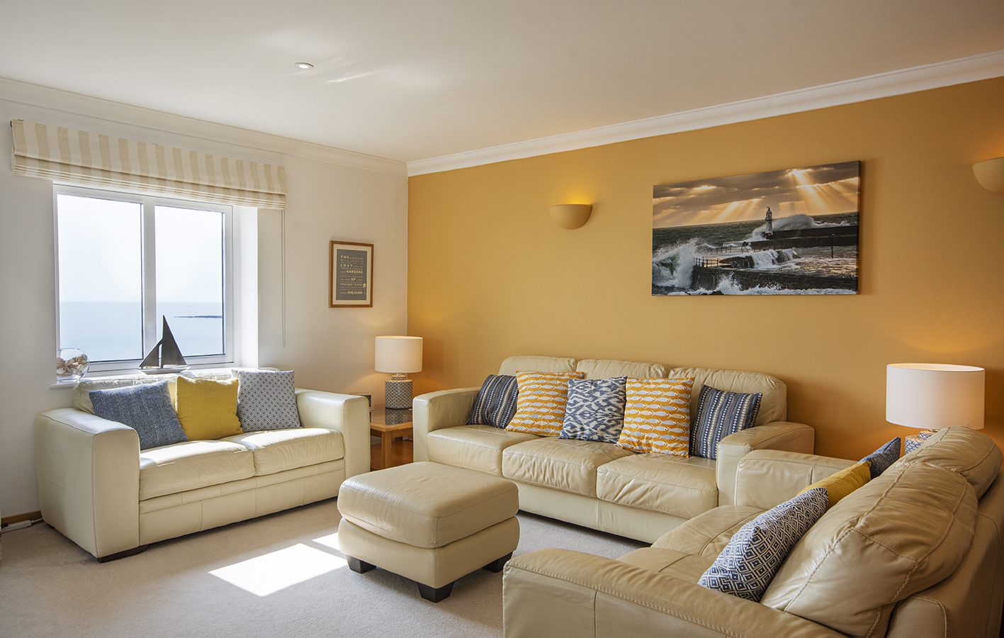 A cozy living room with beige sofas and a matching ottoman arranged around a central point. The room features a warm yellow accent wall, large window with a marine view, and nautical-themed decor, including a sailboat model and ocean artwork.