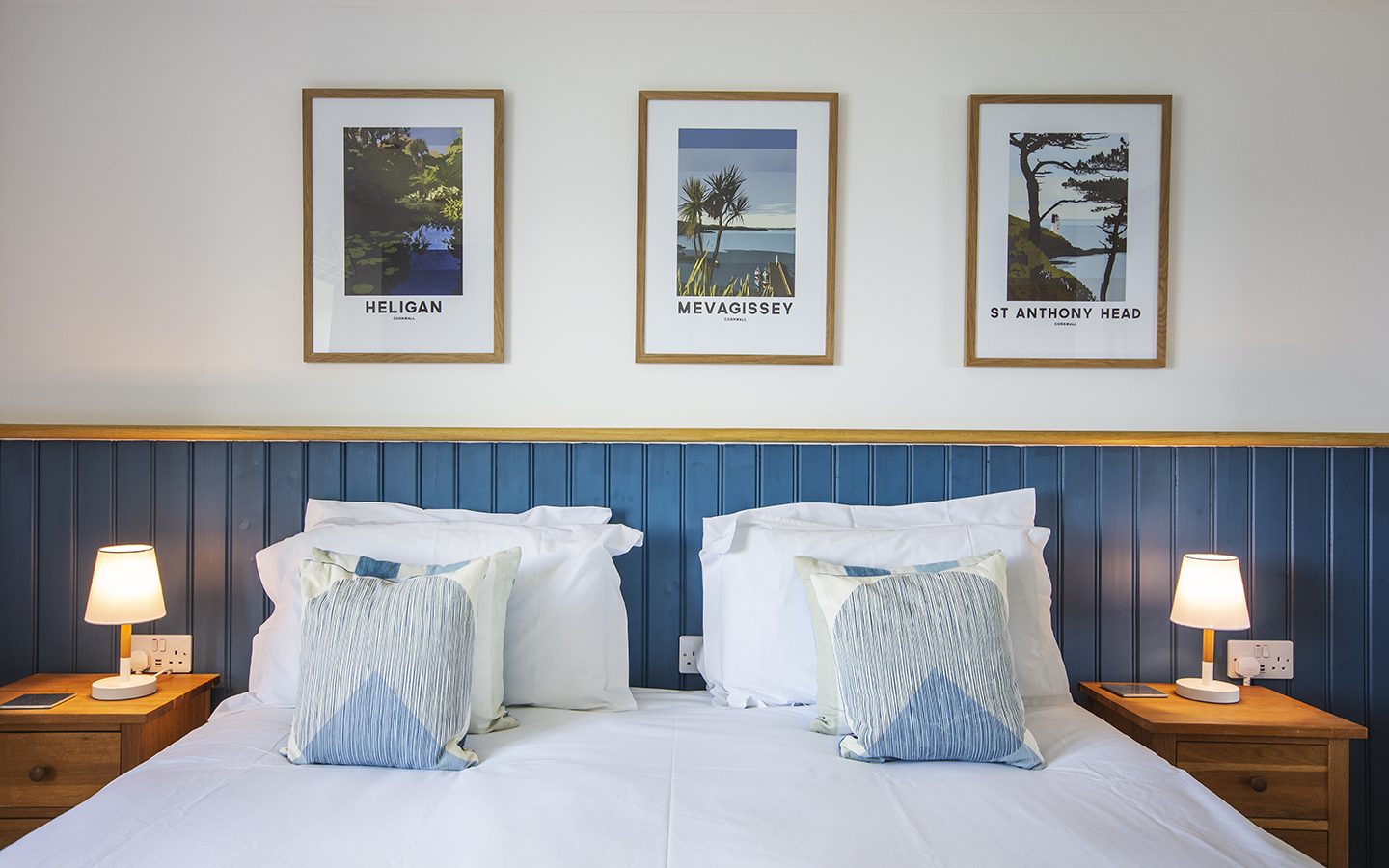 A cozy bedroom features a double bed adorned with two patterned pillows and a blue headboard. Flanking the bed are wooden nightstands with small lamps. Above, three framed landscape posters, including one labeled Marine Point, complement those of Heligan and Mevagissey.