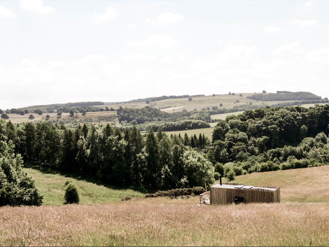 A wooden cabin, perfect for secluded getaways in the UK, sits amidst a grassy field with rolling hills and trees in the background under a partly cloudy sky.