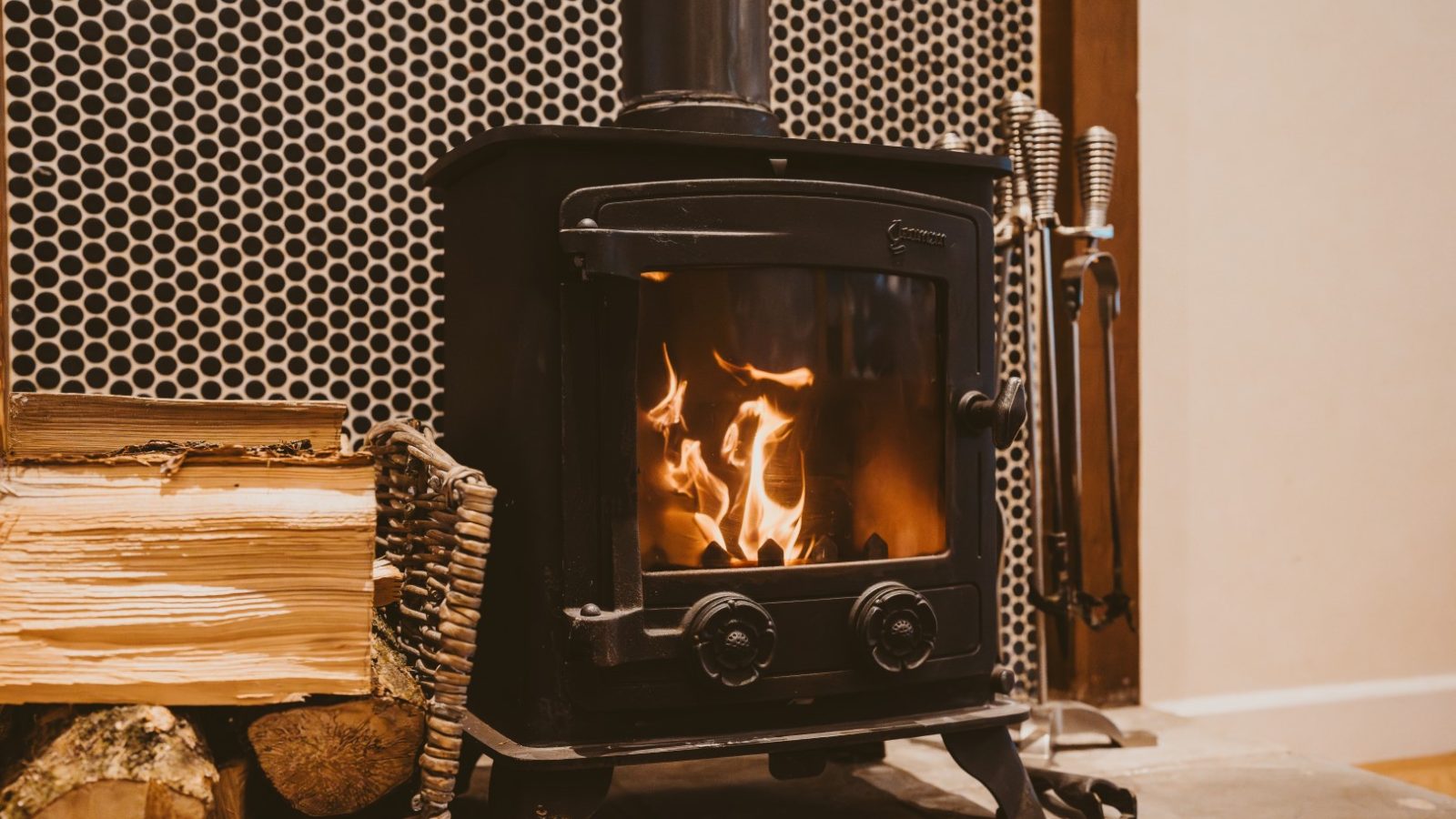 Ploony Holidays Wales: A wood-burning stove with visible flames, surrounded by firewood and tools against a patterned wall.