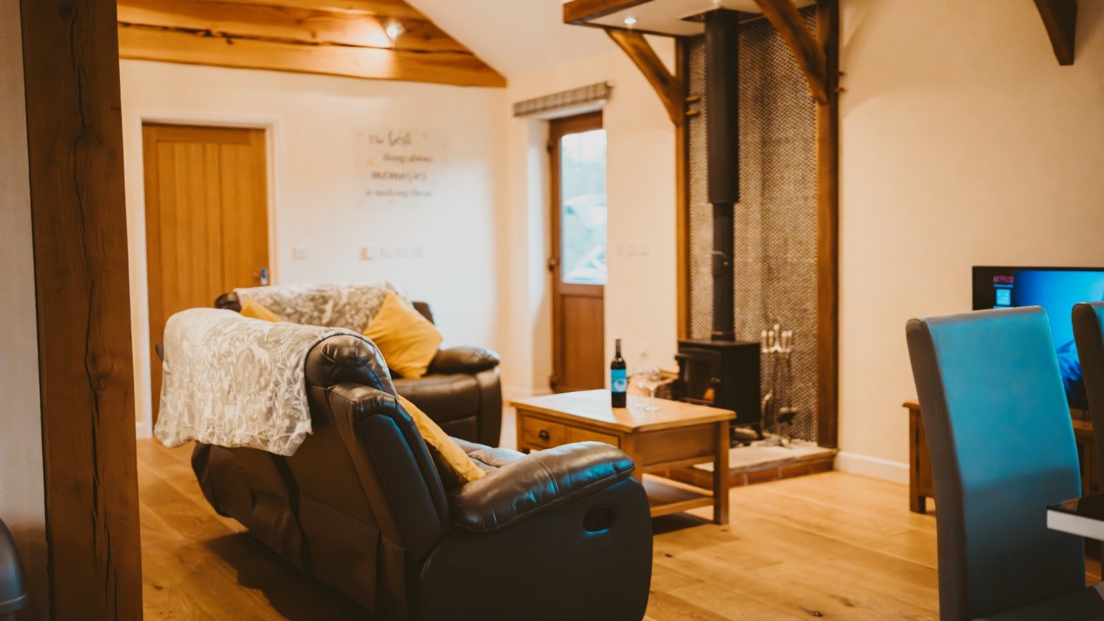 Cosy living room at Ploony Holidays Wales with a wood-burning stove, leather sofas, a wooden coffee table, and wine. Warm ceiling beams.