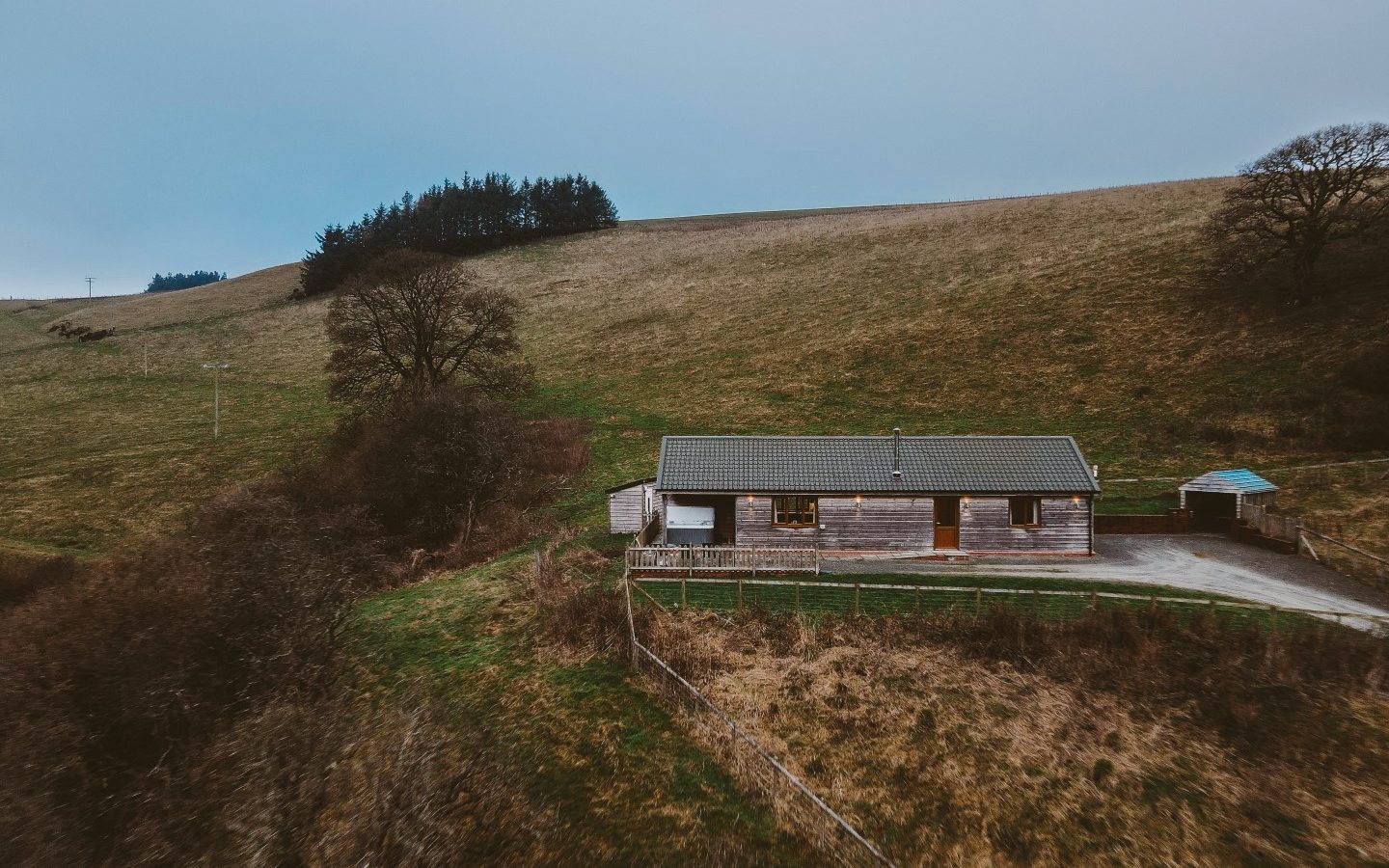 A wooden house with a gray roof sits on a grassy hill in Ploony Holidays, Wales, under a cloudy sky.