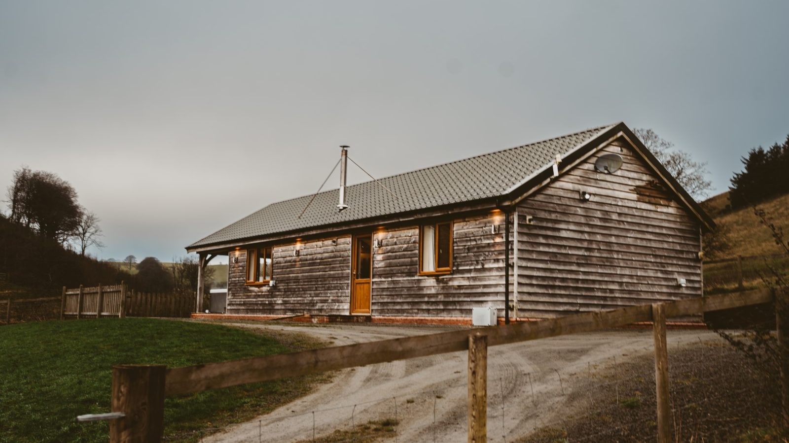 A single-story wooden cabin with a gray roof stands along a dirt path, perfect for Ploony Holidays in Wales.