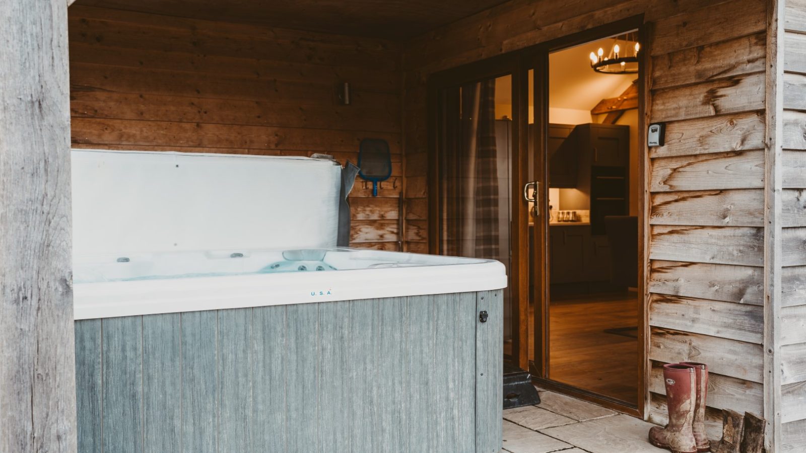 A hot tub on a covered patio, perfect for Ploony Holidays Wales, with wooden walls and boots by the glass door to indoors.