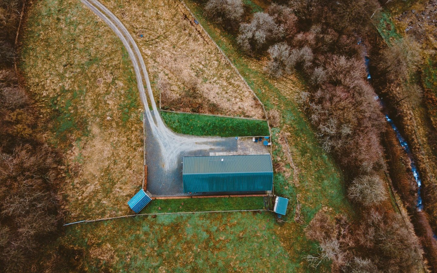 Aerial view of a Ploony Holidays Wales green-roofed building, nestled in fields and trees with a gravel road leading to it.