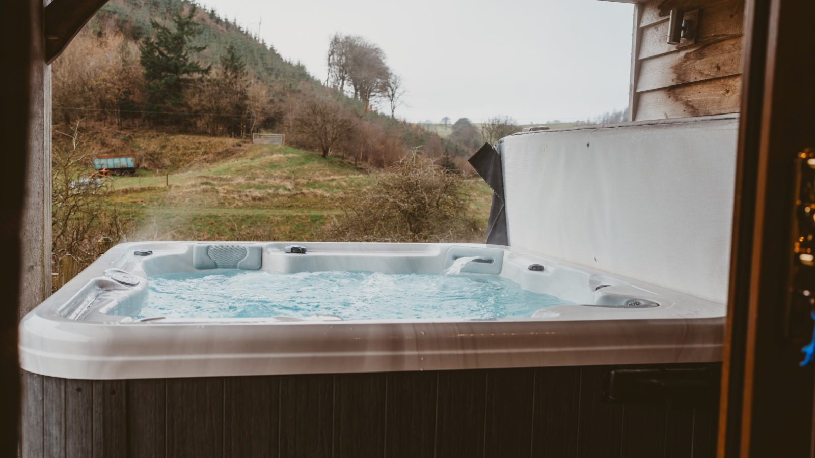 Experience Ploony Holidays Wales in an outdoor hot tub, bubbling under a wooden shelter with scenic views of lush hills and trees.