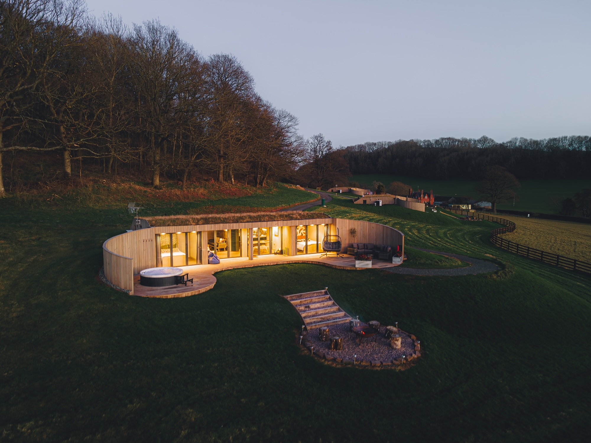 A modern, circular house with large windows and a curved patio is nestled on a grassy hillside, surrounded by trees—one of the best places to go for an October half-term UK escape.