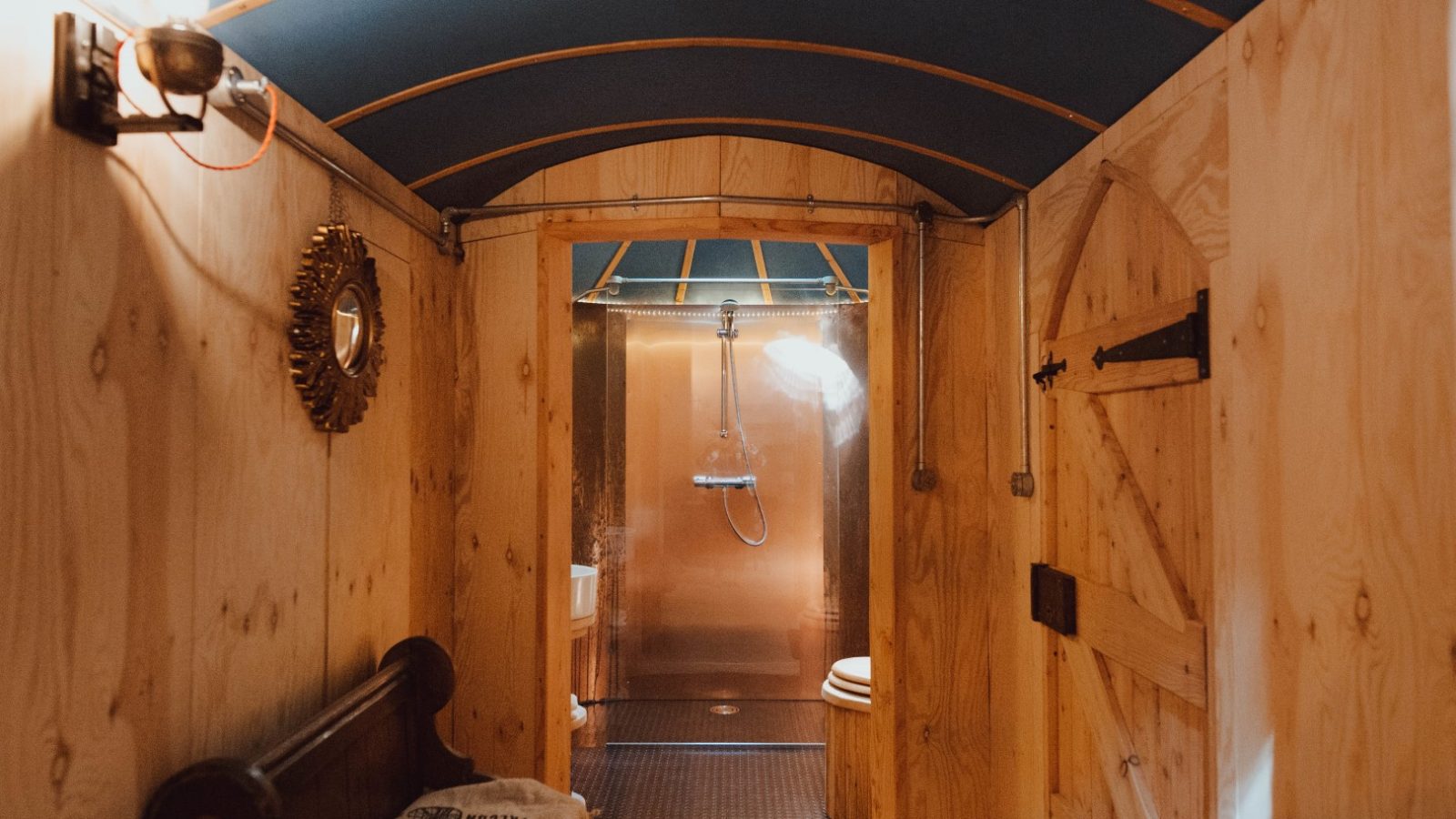 A cozy wooden bathroom in Little Menherion features a curved ceiling, a small round mirror on the left wall, and a rustic door on the right. Steam rises from a sleek modern shower in the back, contrasting with the warm wooden interior design.