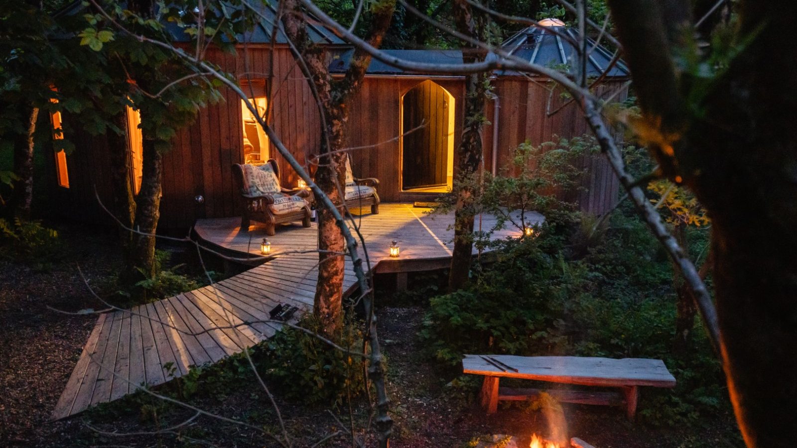 A cozy cabin in the forested enclave of Little Menherion, where warm light glows from the windows. Wooden decks and paths lead to the entrance. A lit fire pit with a bench is in the foreground, surrounded by trees and foliage, creating a serene atmosphere.