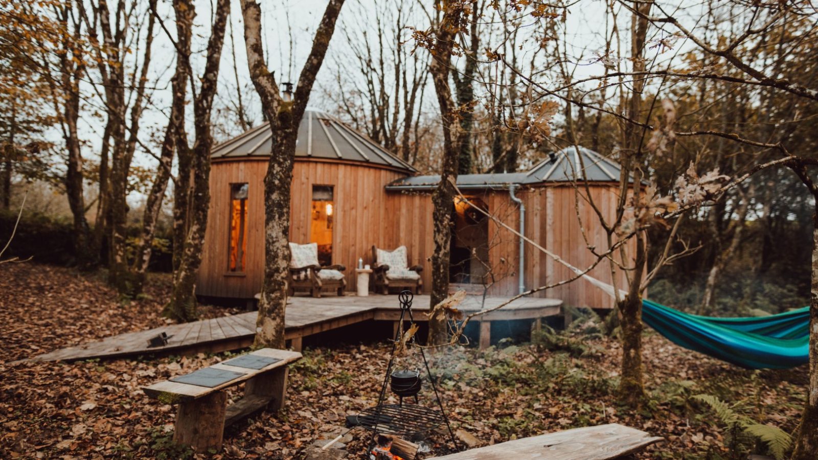 Little Menherion offers a cosy wooden cabin with warm lights nestled among leafless trees in an autumn forest. A hammock sways gently between trees, chairs line the deck, and benches encircle a small fire pit with a pot hanging over it. Fallen leaves blanket the ground in this serene retreat.