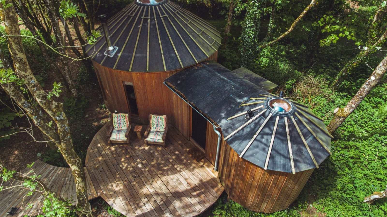 Aerial view of Little Menherion, a unique round wooden cabin with a domed roof, nestled among trees. It features a small deck with two colorful chairs, surrounded by lush greenery, offering a secluded and serene ambiance.