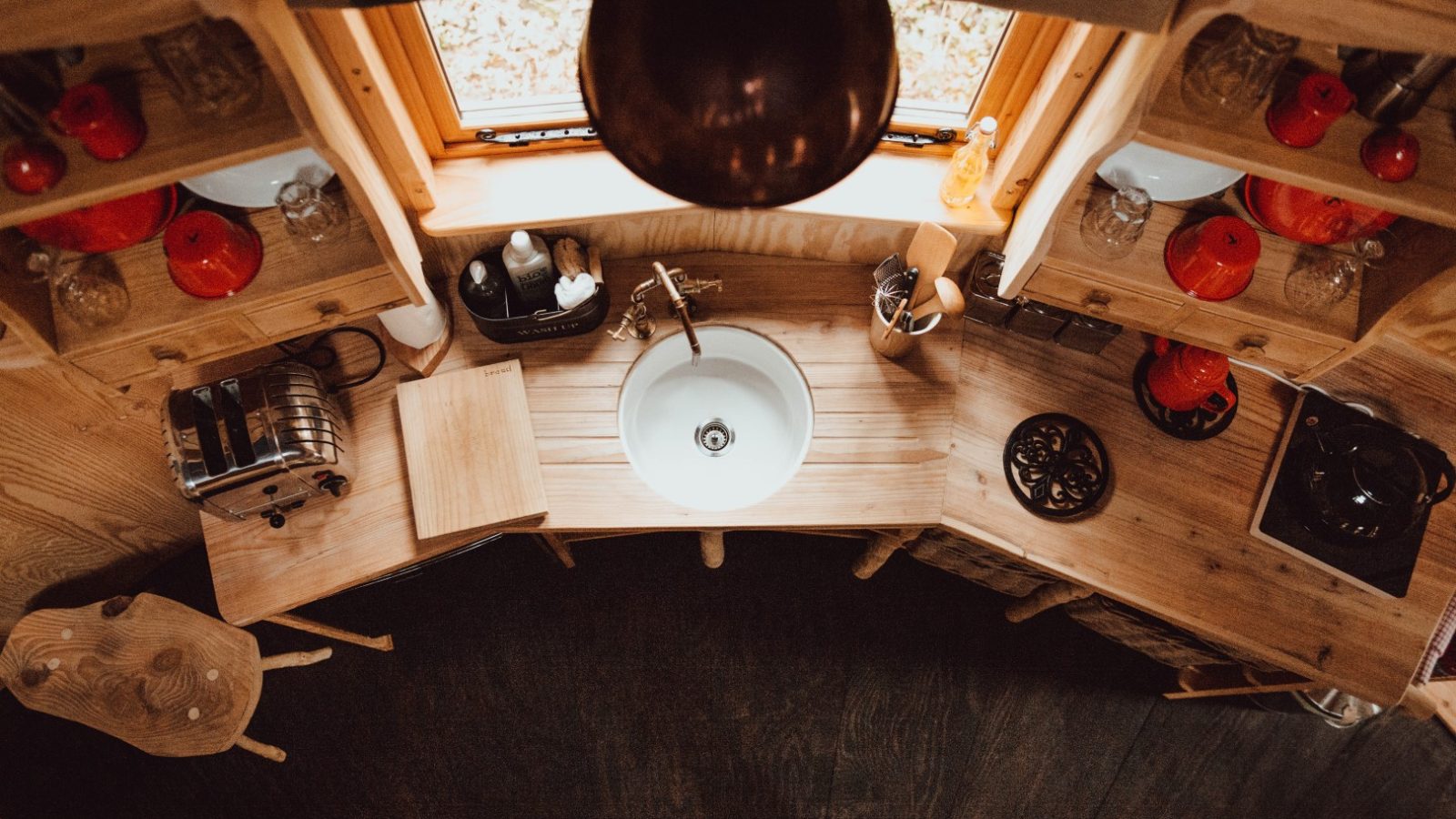 A cozy, rustic kitchen in Little Menherion is viewed from above, featuring a circular white sink, wooden countertops, a toaster, and red kitchenware. Natural light filters in through a window, creating a warm, inviting atmosphere.