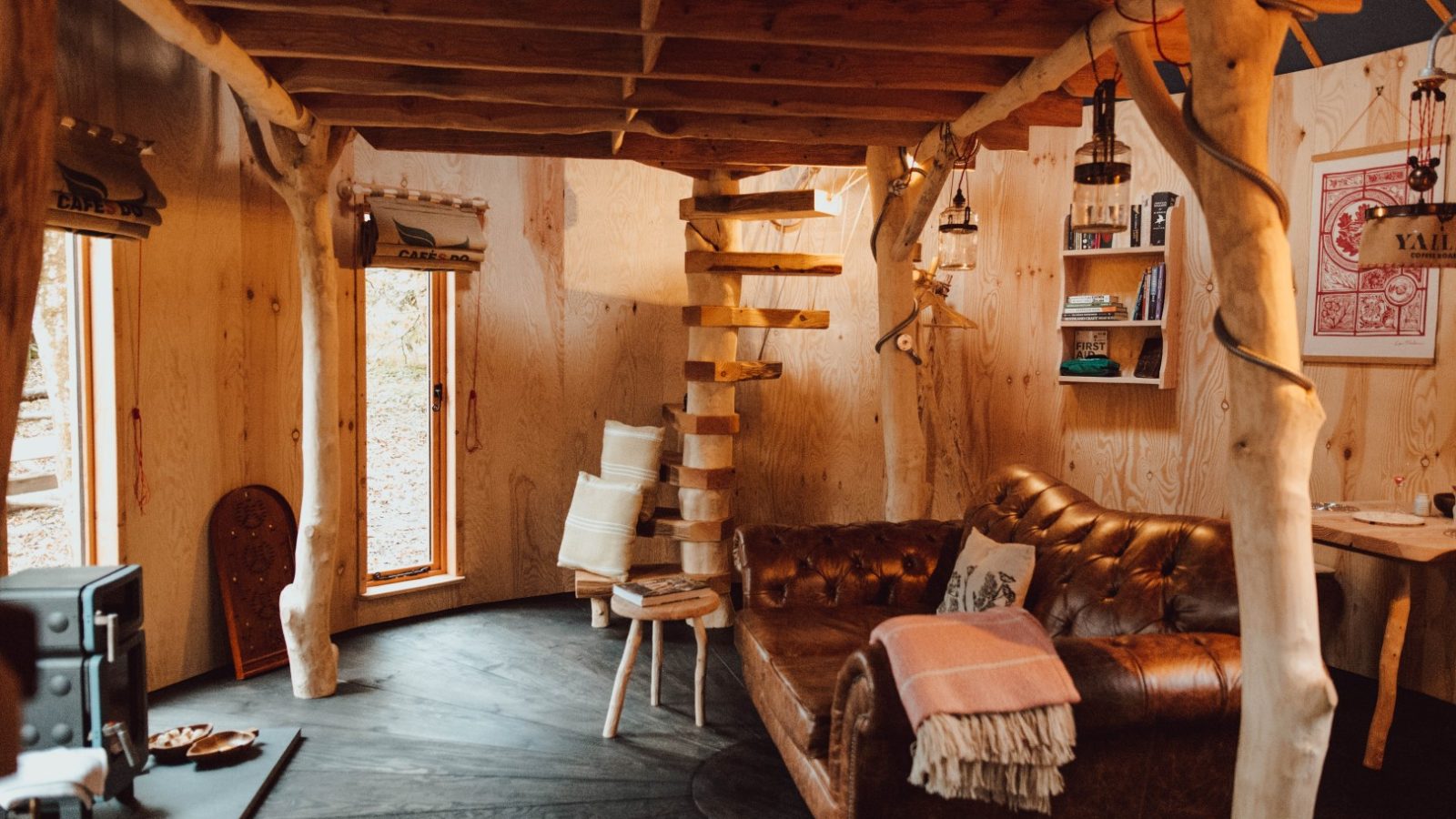 The cozy Little Menherion cabin interior features a wooden spiral staircase, leather sofa, and woodburning stove. Natural wood walls and beams, eclectic decor, and soft lighting merge to create a warm, rustic atmosphere.