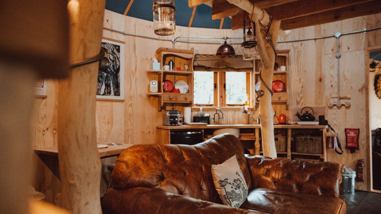 The cozy interior of Little Menherion features a rustic cabin charm with a brown leather sofa, wooden walls, and kitchen shelves. Natural light filters through a small window, illuminating hanging lanterns and various kitchen items. A cushion adorned with a bird design rests on the sofa.