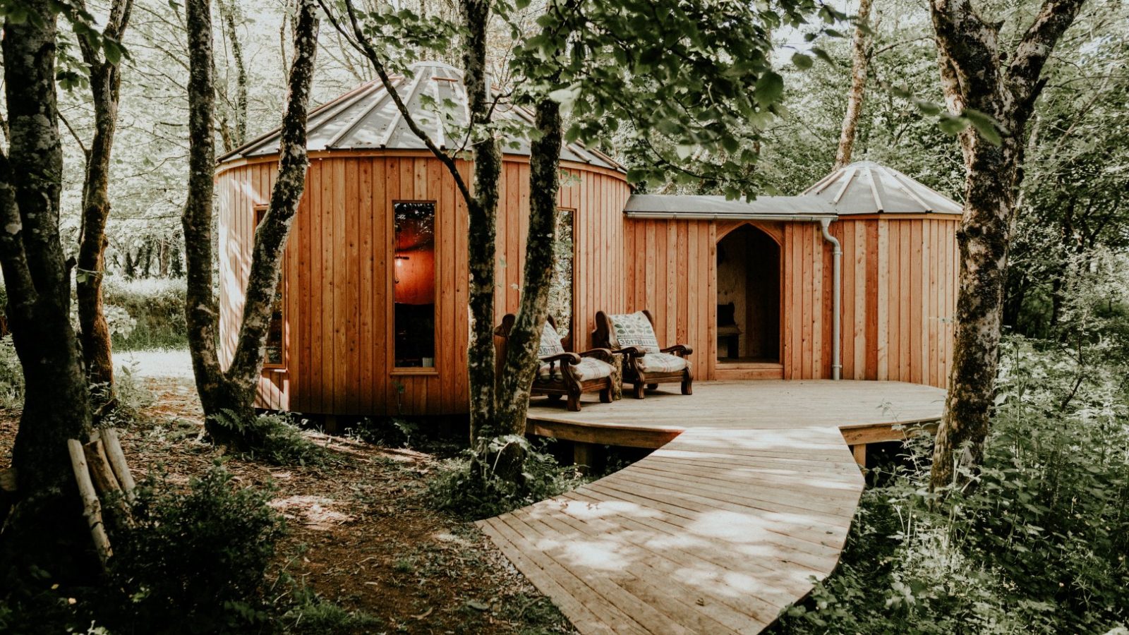 Nestled in the heart of Little Menherion, a rustic wooden yurt awaits amidst lush forest. A charming wooden pathway leads to its entrance, where two chairs on the porch provide a peaceful setting in this verdant haven.