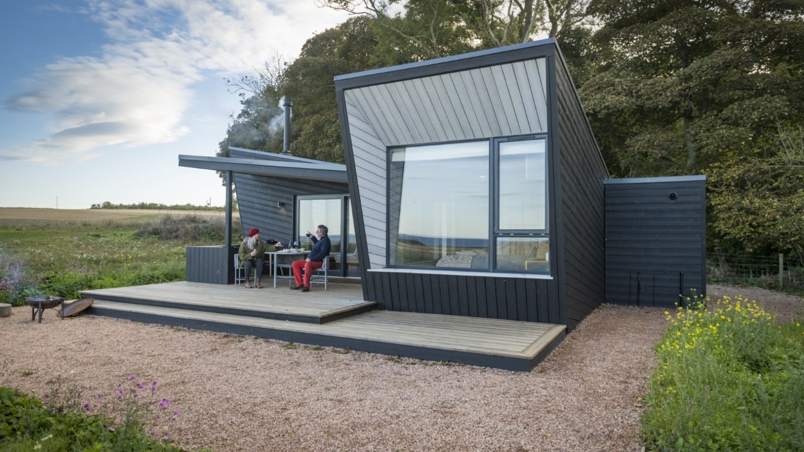 Set amidst the rural beauty of the Cambo Estate, a modern, angular black house with expansive windows offers a serene escape. On its wooden deck, two people relax—one in a red hat and the other in a blue jacket—savoring drinks. Green fields stretch out under the partly cloudy sky, embodying off-grid travel bliss.