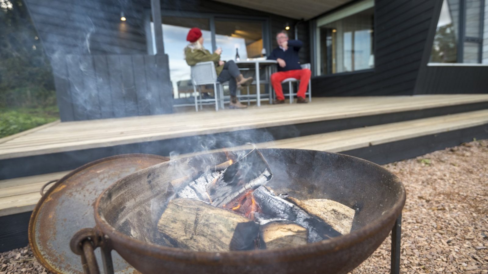 A close-up of a metal fire pit with burning wood and smoke rising. In the background, two people sit on a porch with modern architecture at Cambo Estate, engaged in conversation. One wears a red hat, and the other has red pants. The setting appears relaxed and outdoorsy.