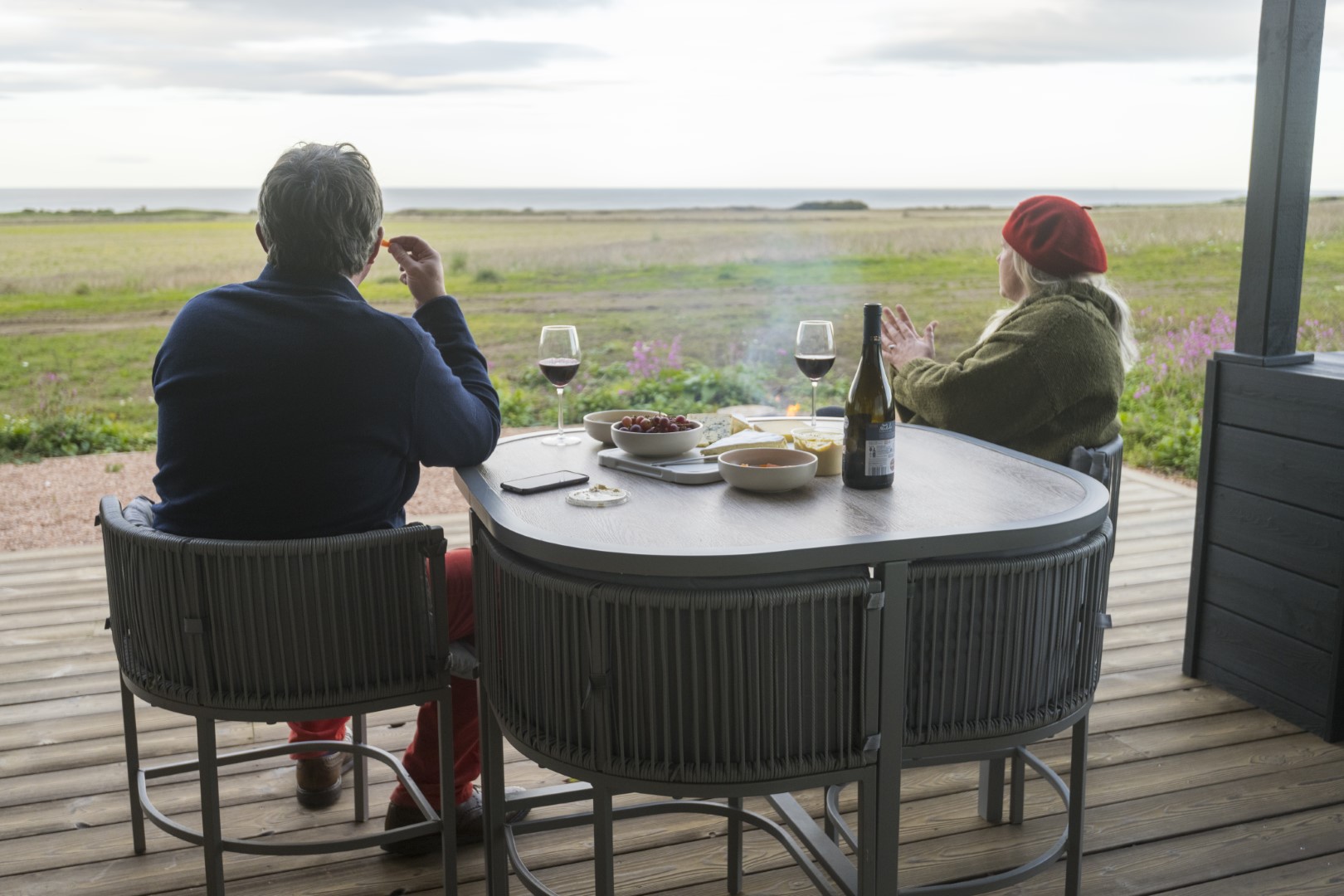 Two people sit at a table on a deck, savoring drinks and snacks, overlooking a vast grassy field and cloudy sky—a perfect scene for those seeking holiday lets that embrace local partnerships.