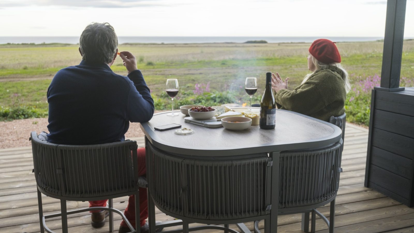 Nestled in the serenity of Cambo Estate, two people relax on a deck overlooking a scenic field and ocean beyond. On the table, glasses of red wine accompany snacks. The overcast sky adds tranquility to this off-grid travel escape, while flowers gently color the horizon.