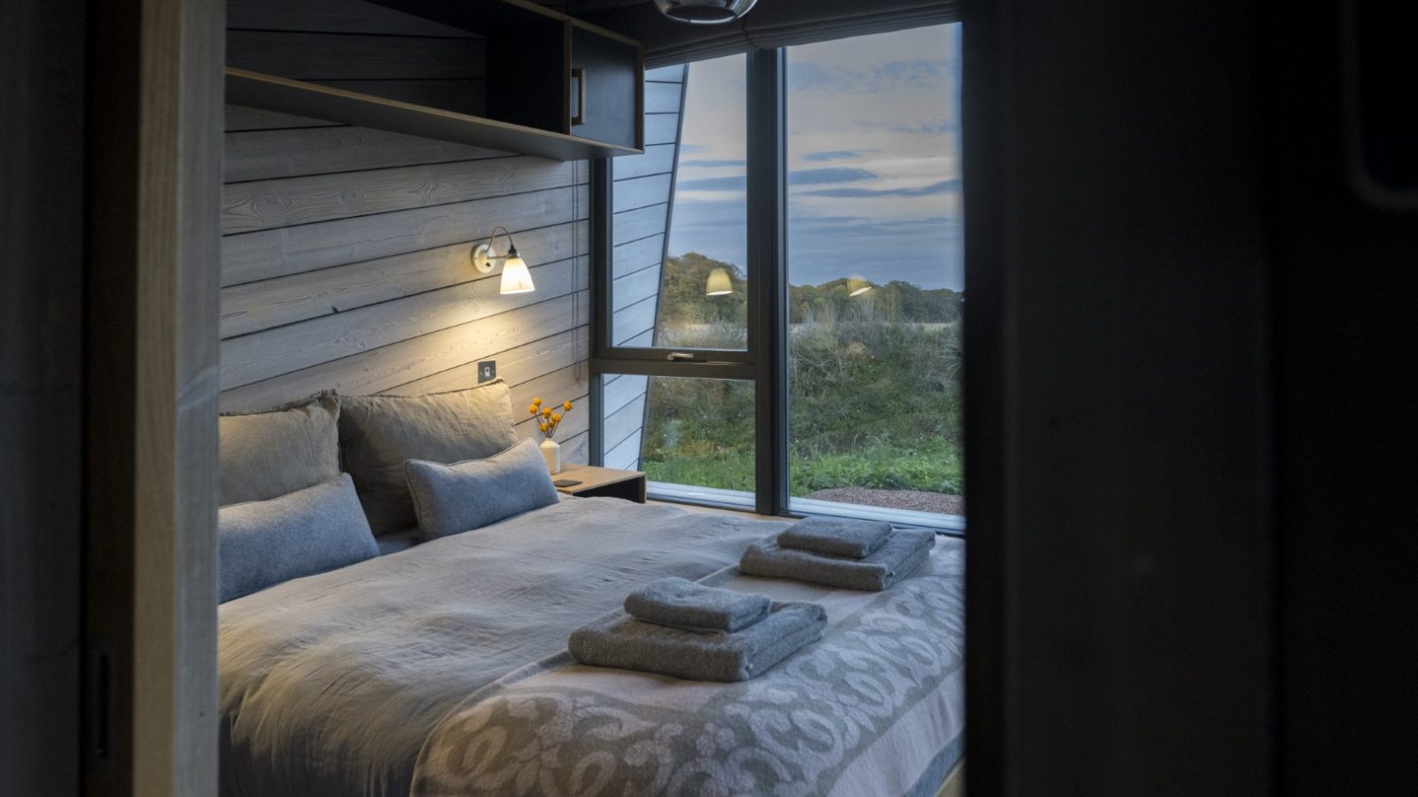 A cozy bedroom at Cambo Estate features a neatly made bed with folded towels on top. Soft lighting and large windows provide a view of the serene, off-grid landscape with hills. Wood paneling adds warmth, creating a relaxing atmosphere.