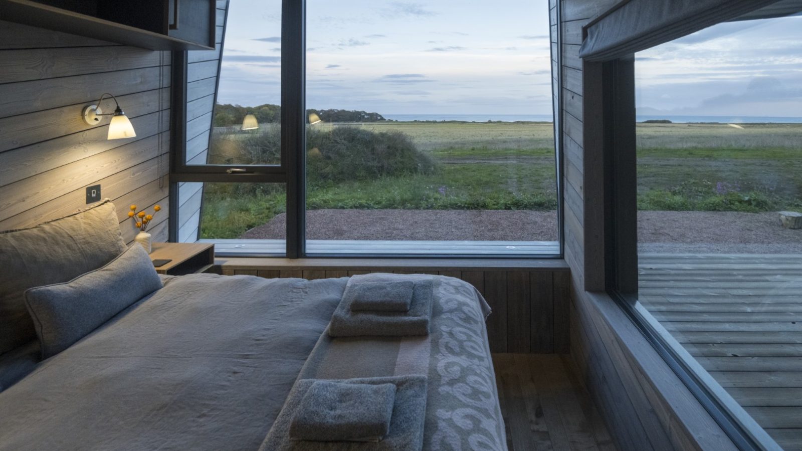 A cozy bedroom at Cambo Estate with large windows overlooks a serene landscape of grass and open sky. The room features wooden walls, a plush bed with folded towels, and warm lighting from a wall-mounted lamp, creating a tranquil atmosphere perfect for an off-grid travel experience.