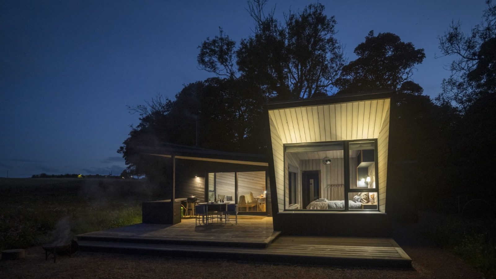 Nestled in the serene Cambo Estate, this modern cabin at night boasts large windows illuminated from within. The angular design and wooden deck with outdoor seating enhance its charm. Surrounded by trees and a dark sky, it offers a cozy, off-grid travel experience.