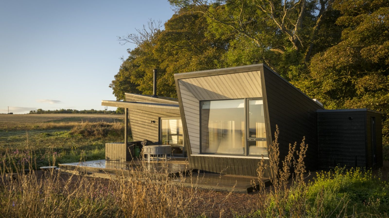 A modern, angular cabin with large windows sits surrounded by trees and fields under a clear sky on the Cambo Estate. The structure features dark wood siding and a deck, creating a contrast with the natural greenery and golden grassland, ideal for off-grid travel enthusiasts.