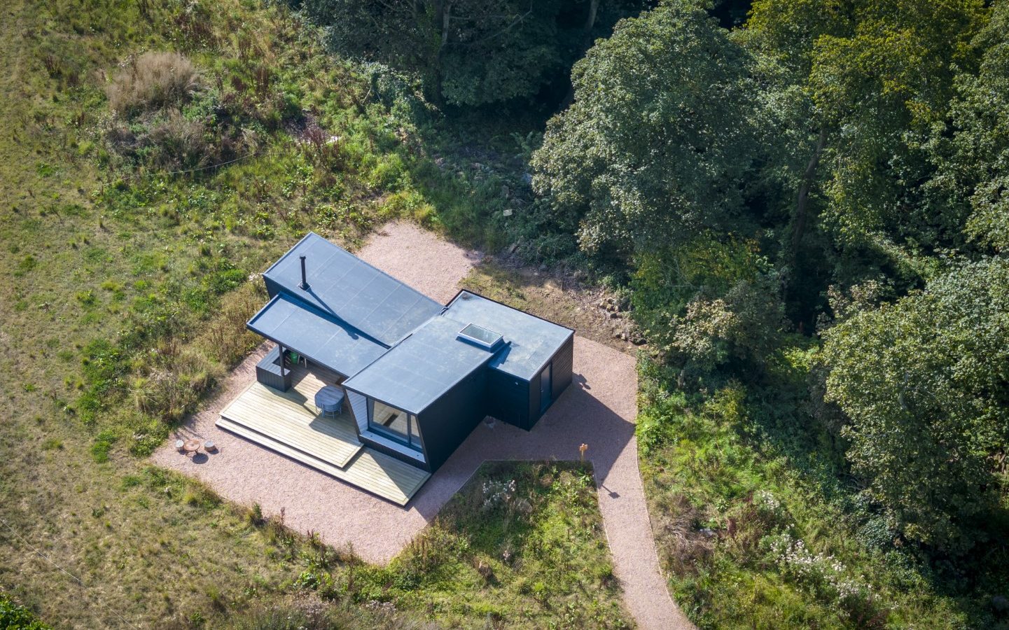 Aerial view of a modern, small black home with a flat roof, nestled in the lush, wooded landscape of Cambo Estate. The house features a wooden deck with outdoor furniture, surrounded by greenery and a winding path leading to the entrance—perfect for off-grid travel enthusiasts.