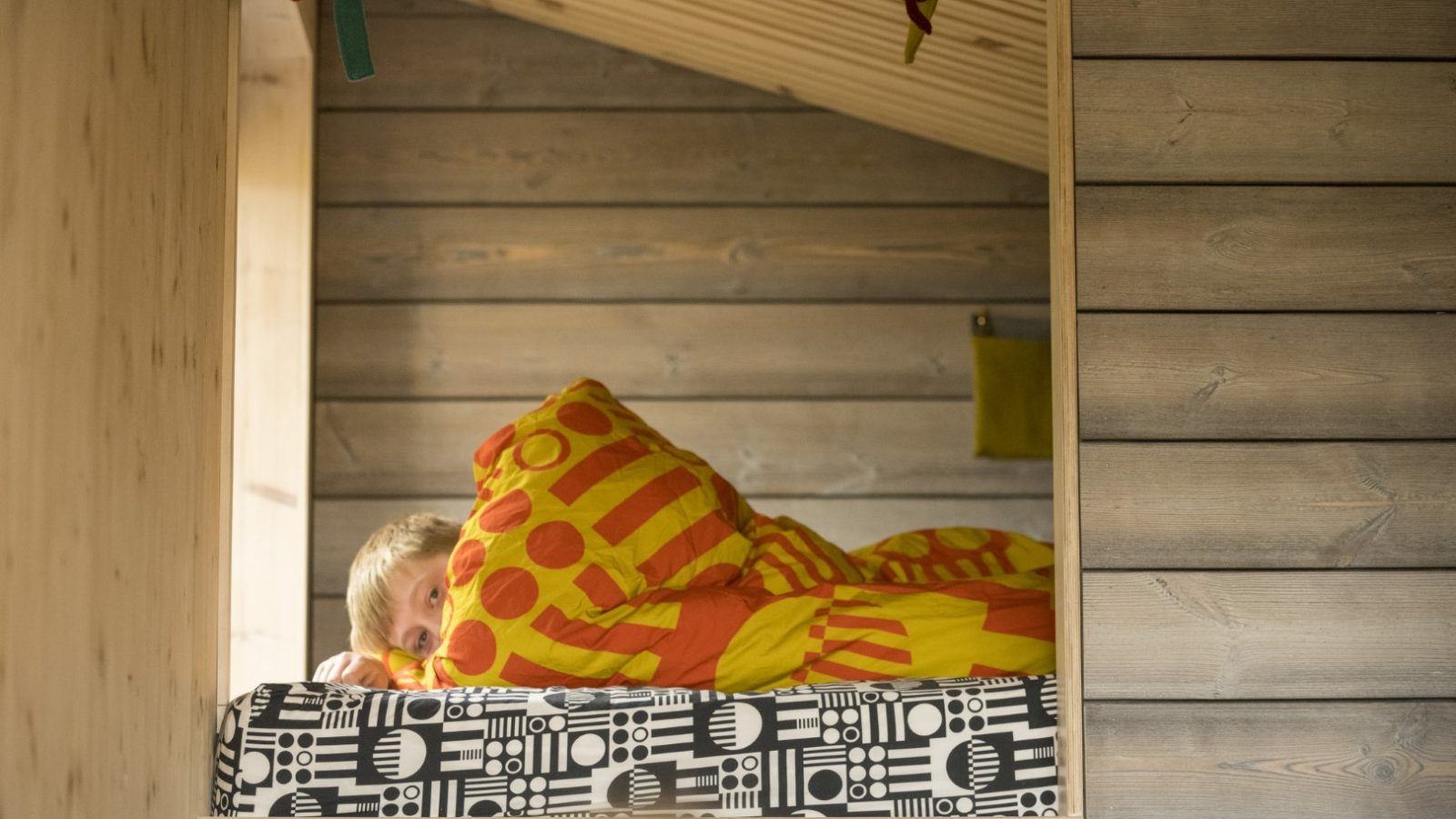 A person lies in a wooden loft bed, wrapped in a bright orange and yellow polka-dot duvet. The cozy space resembles an off-grid travel retreat, with its natural wooden walls and slanted roof. The bed features a patterned black-and-white mattress, creating a charming escape akin to Cambo Estate.