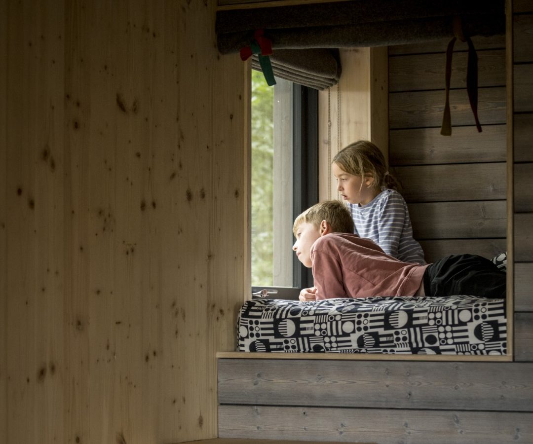 Two children gaze out a window from a cozy wooden loft bed at the Cambo Estate. The room features wood paneling and built-in stairs leading up to the bed, creating a rustic and warm atmosphere perfect for off-grid travel enthusiasts seeking tranquility.