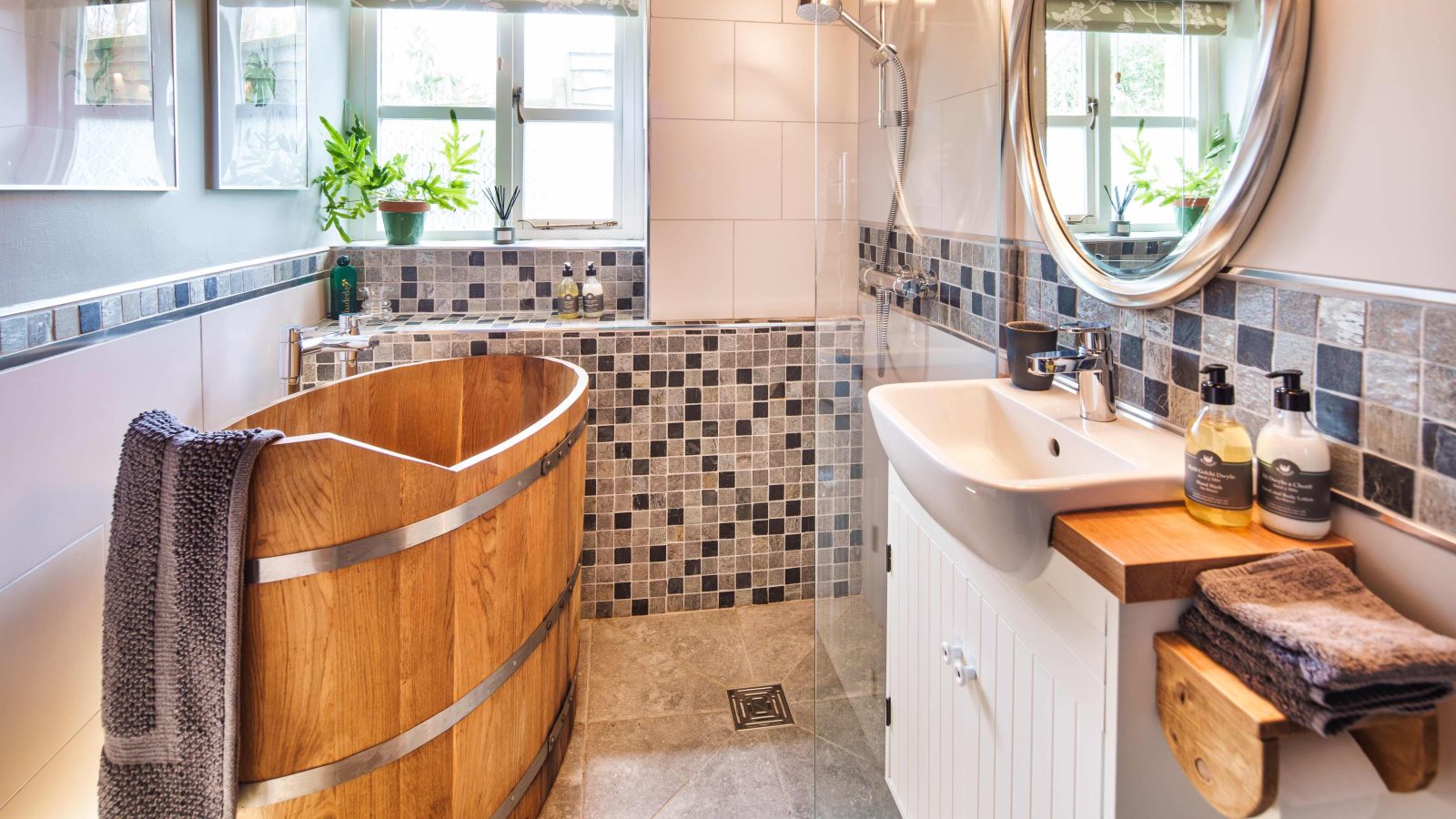 Nestled within the charming Glanmedeni accommodation, this cozy bathroom features a wooden barrel bathtub, a glass shower, and a white sink with a round mirror above. Gray and white tiles adorn the walls, while plants on the windowsill add a refreshing touch of greenery.