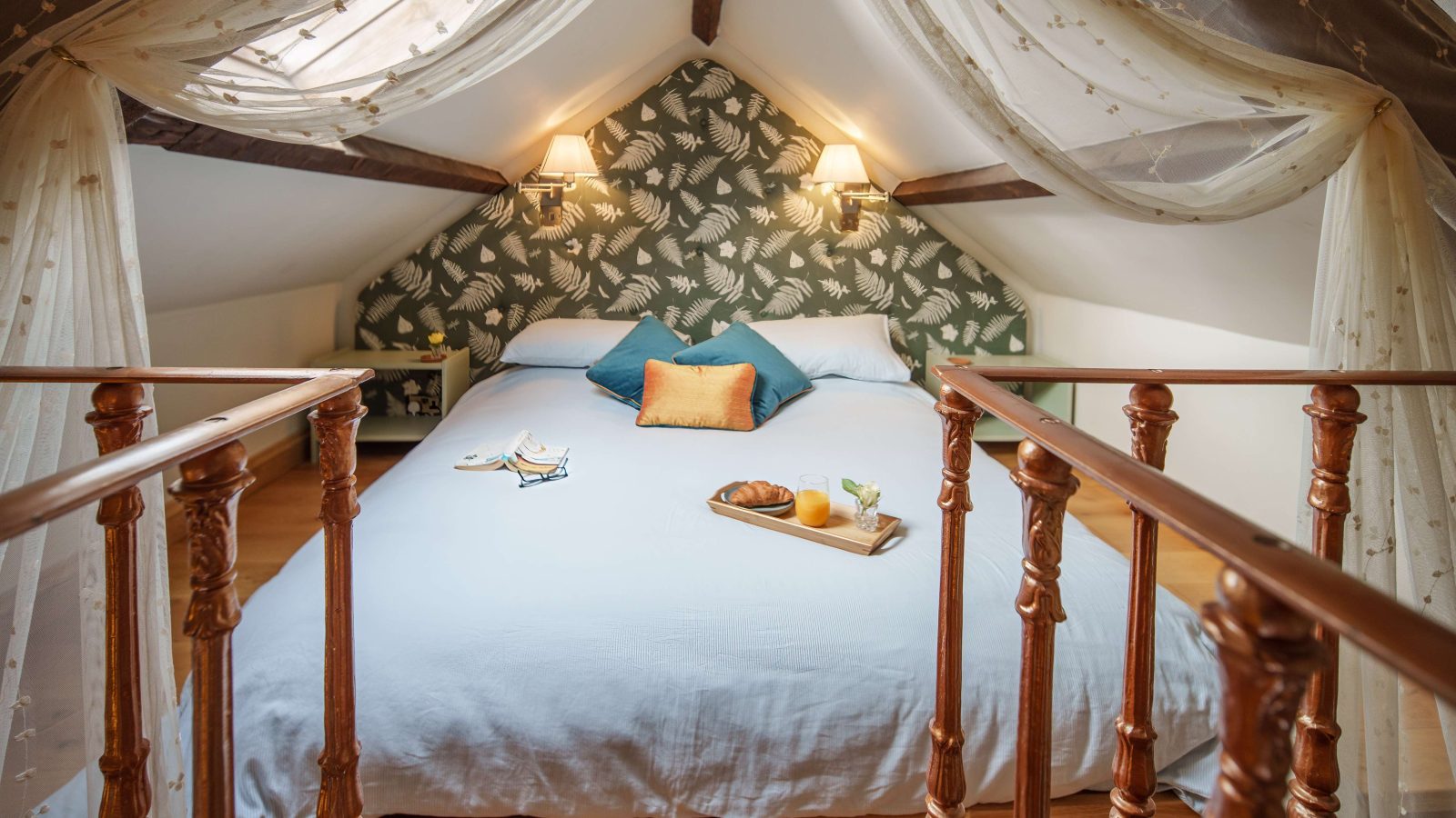 Cosy attic bedroom at the Forge at Glanmedeni, featuring a double bed adorned with blue and orange pillows. A tray with breakfast items is placed on the bed. Soft curtains hang from the ceiling, and light filters through a skylight, illuminating the room.