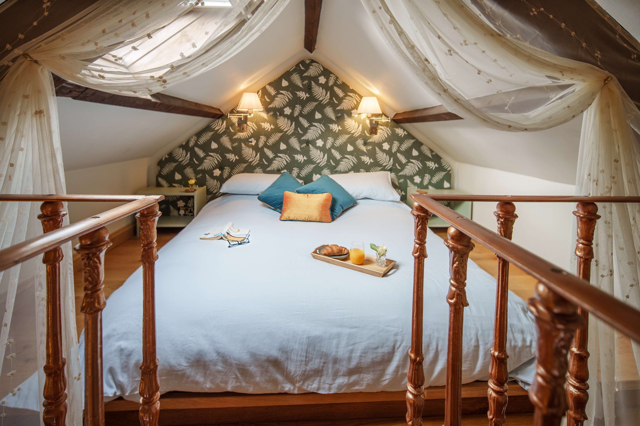 Cosy attic bedroom at the Forge at Glanmedeni, featuring a double bed adorned with blue and orange pillows. A tray with breakfast items is placed on the bed. Soft curtains hang from the ceiling, and light filters through a skylight, illuminating the room.