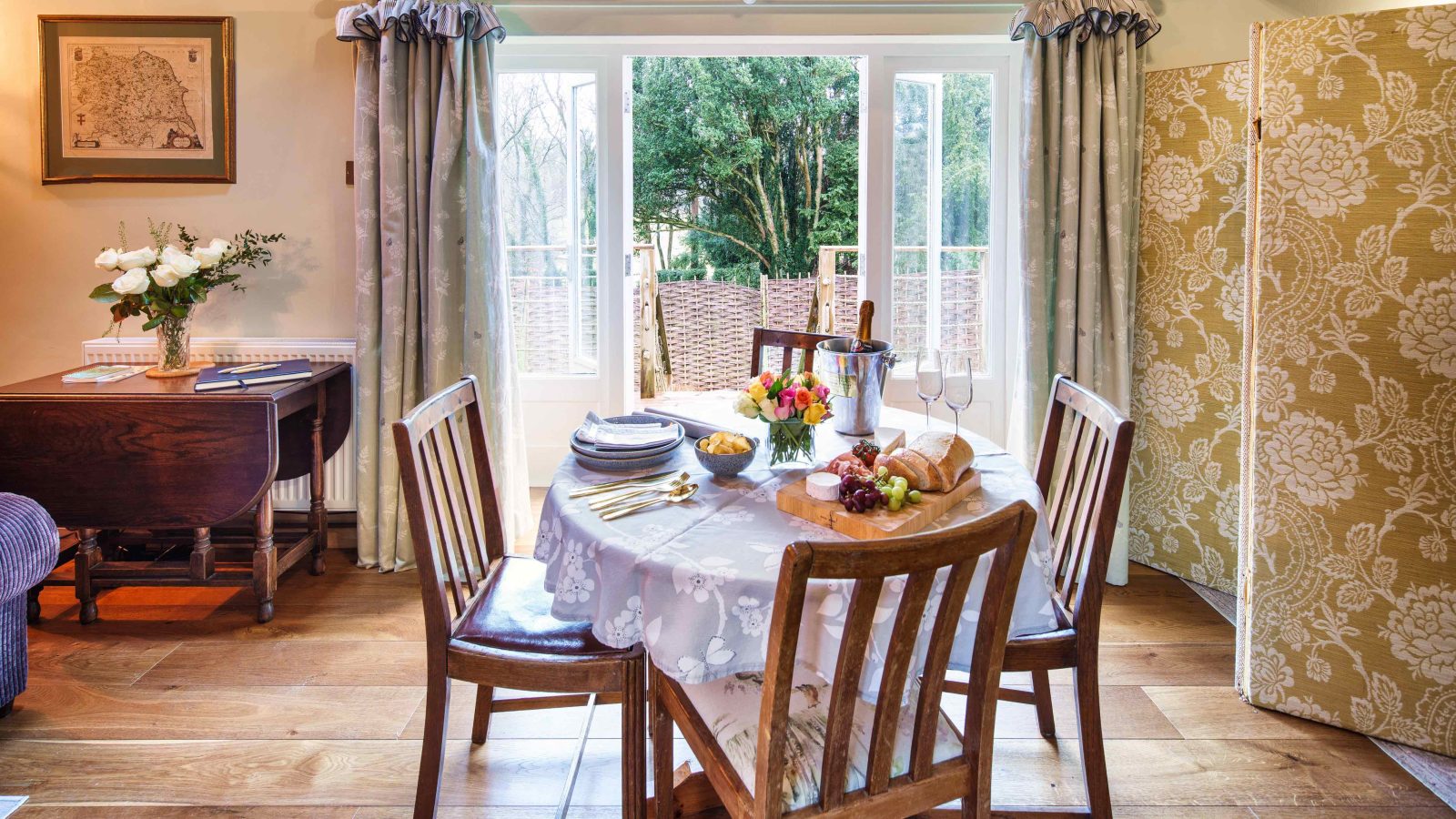 The Glanmedeni dining table set, perfect for a meal in a cozy room with wooden floors, features a white cloth adorned with plates, cutlery, wine glasses, and a basket of bread. French doors open to a lush garden, while a vase of flowers and The Forge's decorated screen enhance the ambiance.