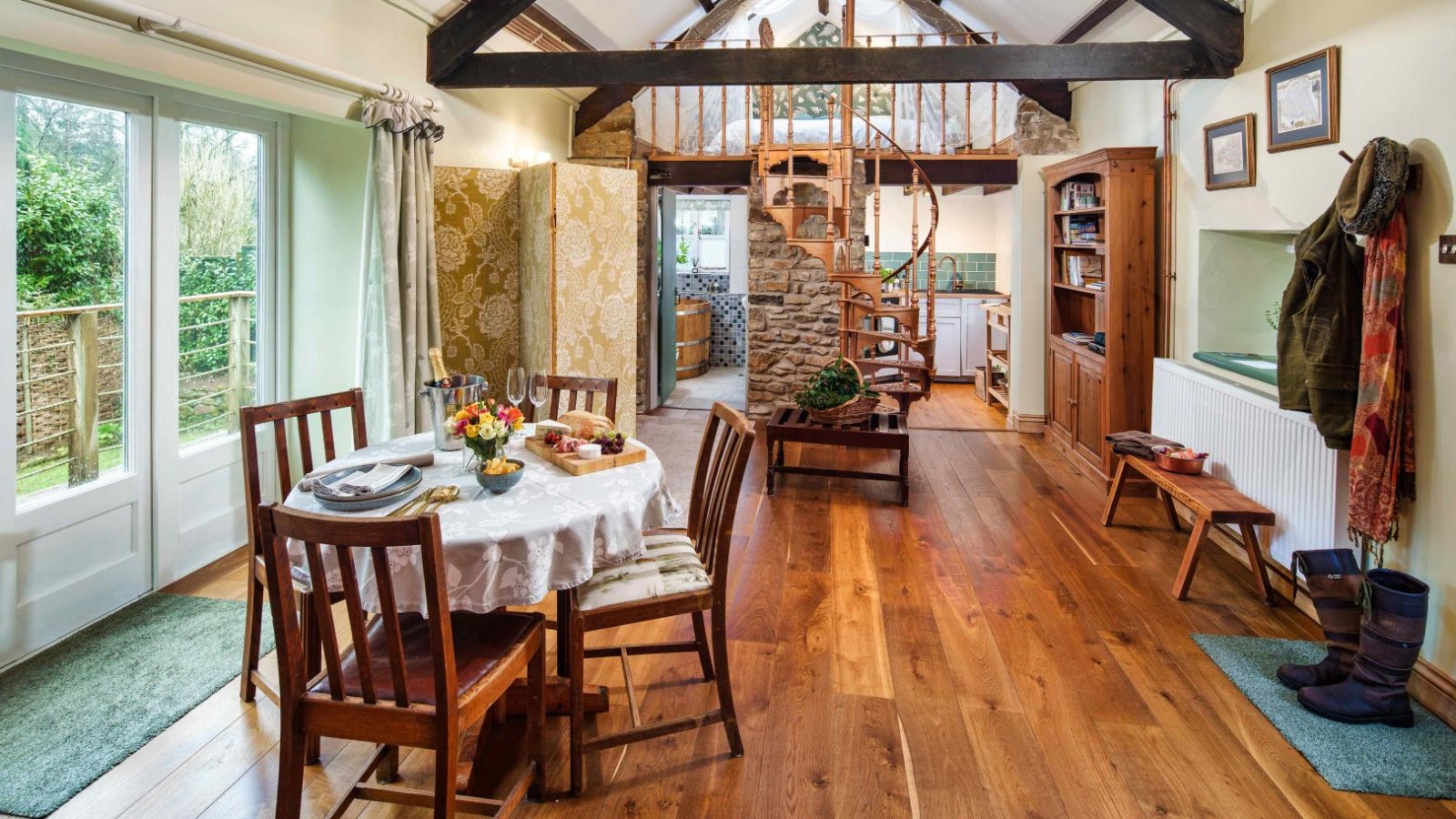 A cozy, rustic living space at Glanmedeni features wooden floors and a spiral staircase leading to a loft. A dining table set for two sits by the small kitchen, with a coat hanging by the radiator. Large windows let in natural light, revealing The Forge garden outside.