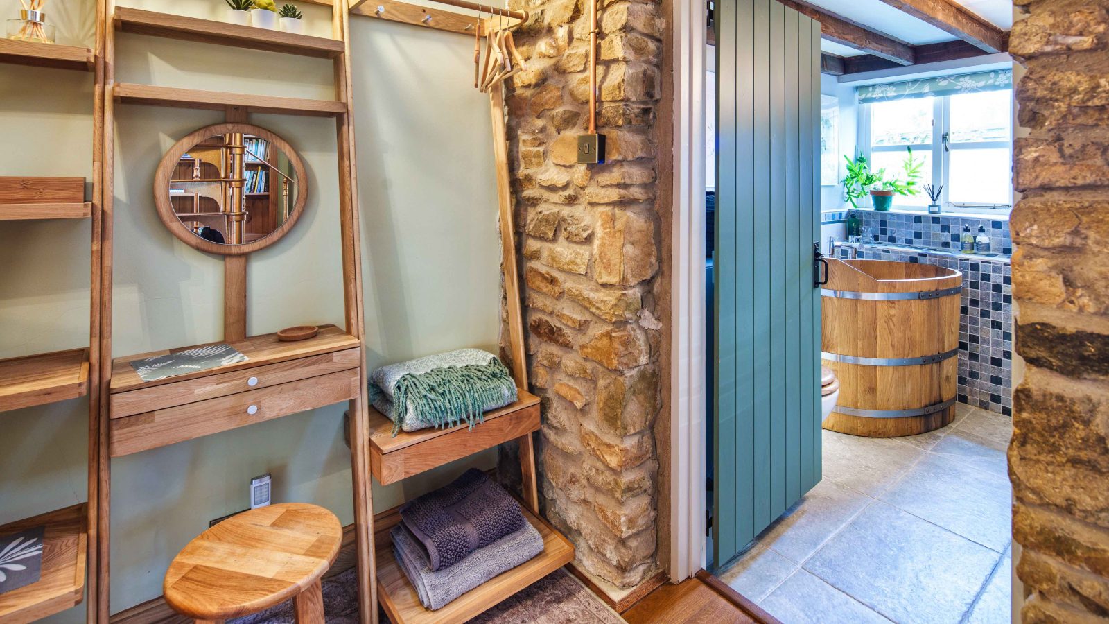 A cozy room with wooden furniture from Glanmedeni features a small stool, shelves with folded towels, a circular mirror, and a stone wall. An open door reveals a bathroom with a Forge-style wooden barrel tub and tile flooring under a bright window.