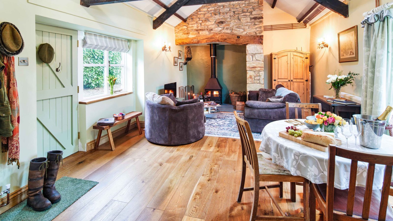 Cozy rustic living and dining room in Glanmedeni with exposed beams, stone fireplace, and wood-burning stove. Two armchairs face the fire. A round dining table is set with flowers and bread. Light streams through a window, highlighting wooden floors. Boots by the door evoke a sense of home at the forge.