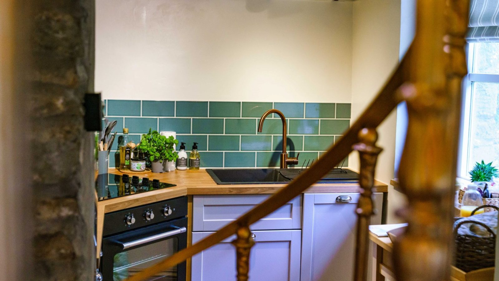 A cozy kitchen with wooden beams and green subway tile backsplash exudes a Glanmedeni charm. It features a black oven, wooden countertops, copper sink faucet, and potted herbs. A golden staircase railing is partially visible in the foreground.