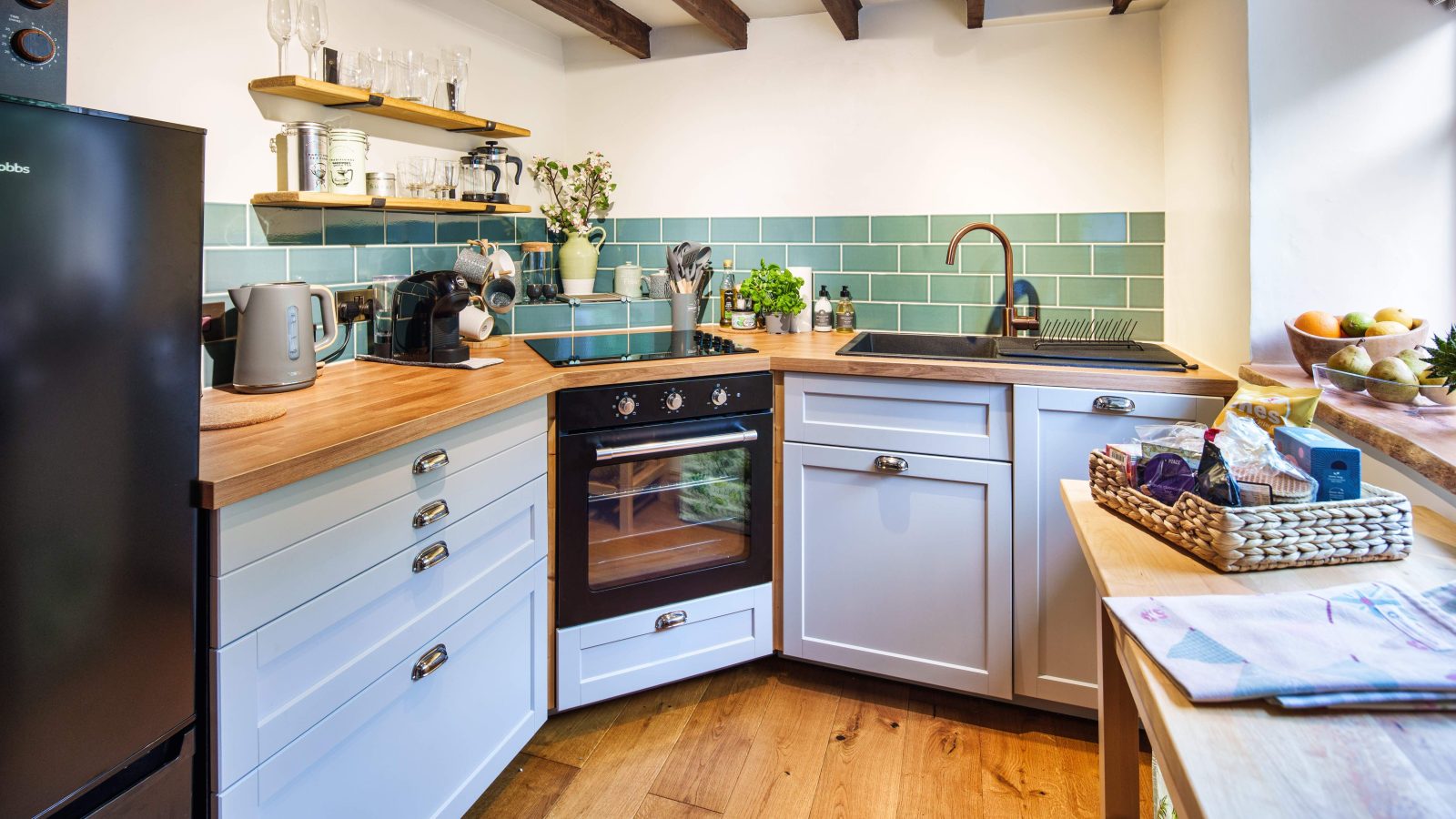 A cozy kitchen with light blue cabinets and wooden countertops reminiscent of The Forge, featuring a teal tile backsplash. The space includes a black oven, sink, and various kitchen appliances. Potted plants from Glanmedeni line the windowsill alongside a vibrant fruit basket.