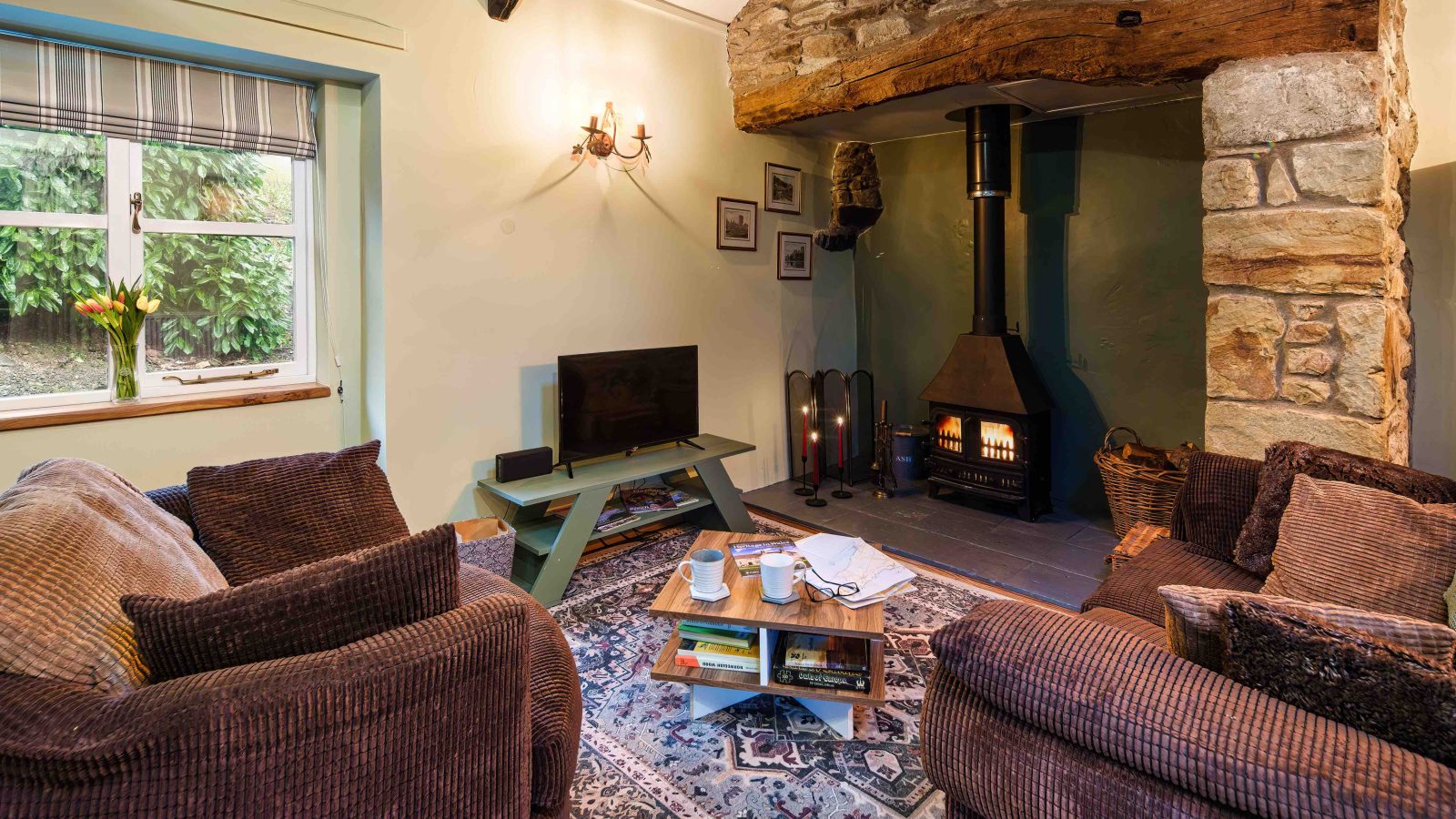 A cozy living room at Glanmedeni features two brown sofas, a wooden coffee table, and a patterned rug. There's a wood-burning stove with a stone surround and a rustic wooden beam. A small TV sits on The Forge green stand by the window, overlooking lush plants outside.