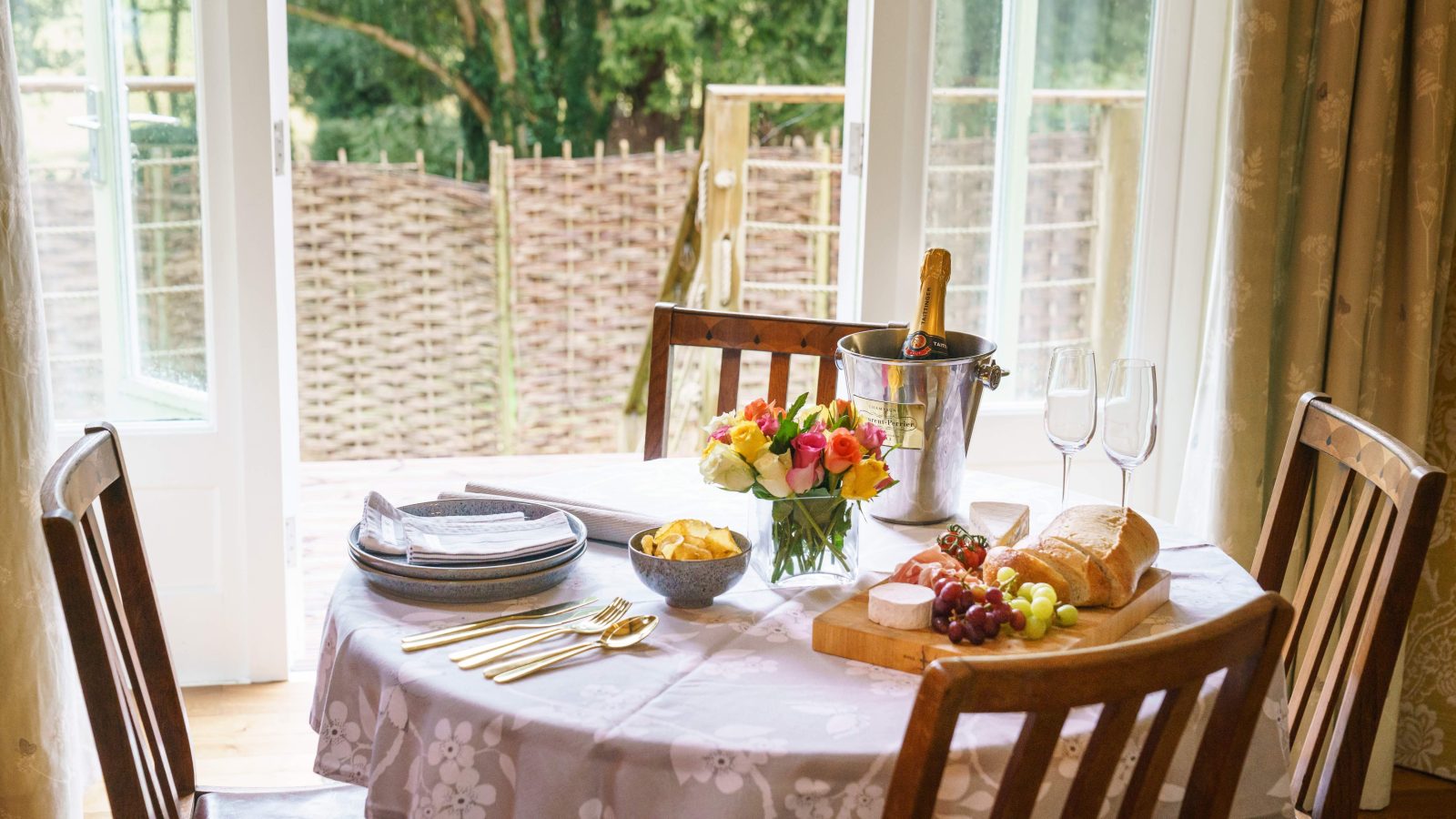 A round dining table with a floral tablecloth is set for a meal at The Forge. It boasts a bouquet of colorful flowers, a breadboard with cheese, grapes, and bread, plates, and a bucket with champagne and two glasses, all perfectly positioned in front of a window.