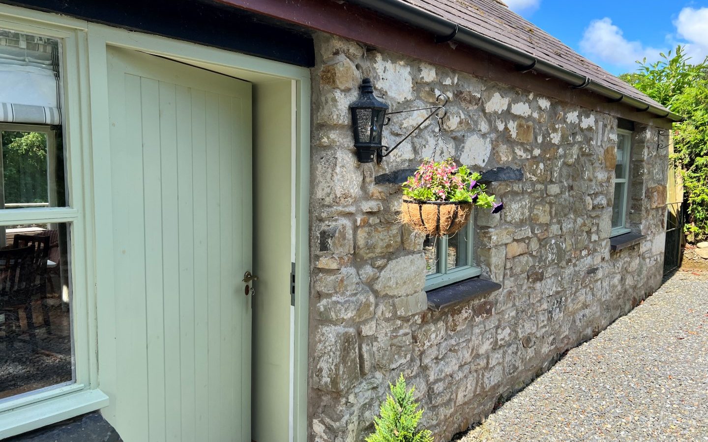 A quaint stone cottage in Glanmedeni features a light green wooden door. Above the door, a hanging basket filled with flowers complements the black lantern on the stone wall. This charming retreat is surrounded by a gravel path under a clear blue sky.