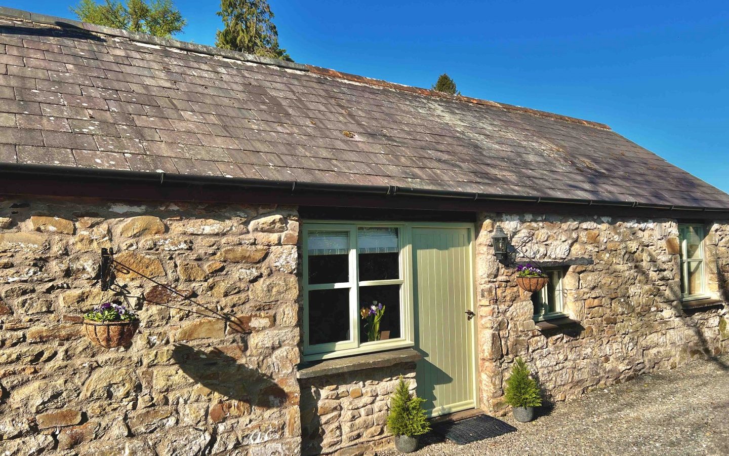 A rustic stone cottage known as The Forge, with a gray slate roof and a pale green door, stands proudly. Two small potted trees by the entrance and hanging flower baskets adorn the walls, painting a charming scene against the clear blue sky.