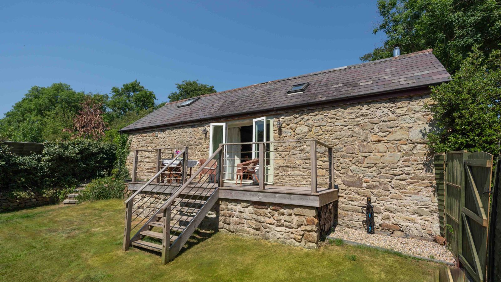 A rustic stone cottage, known as Glanmedeni, with a slate roof and wooden deck sits in a grassy yard. The deck, reminiscent of a forge's charm, features chairs and a small table. Trees and bushes surround the property under a clear blue sky.