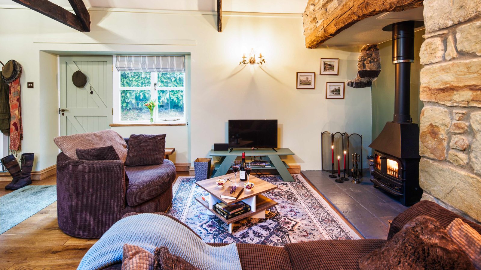 A cozy living room at Glanmedeni with exposed wooden beams, a stone fireplace, and a TV on a wooden stand. The room features plush brown sofas, a patterned rug, and a coffee table with wine and glasses. A door and window reveal lush greenery outside.