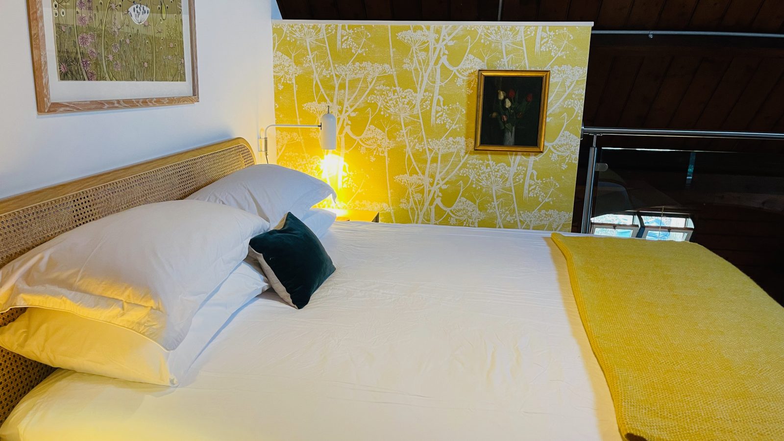 A cozy bedroom with a neatly made bed featuring white linens and decorative pillows. The room's wooden ceiling and yellow accent wall with floral patterns embody a mission-style charm. Two framed pictures hang on the wall, enhancing the warm and inviting atmosphere.