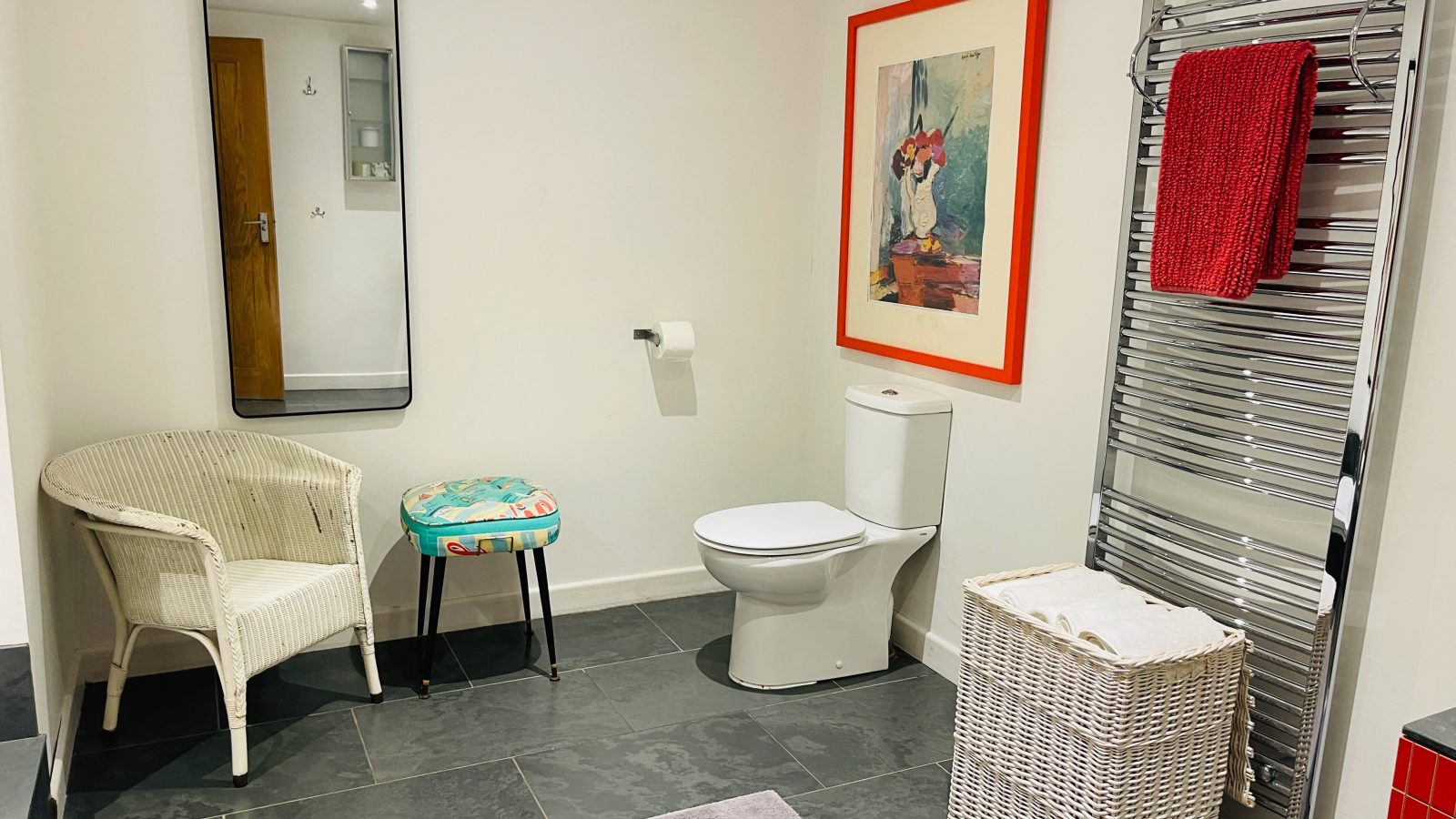 A modern bathroom styled with a wicker chair and colorful cushion, featuring The Mission's charm. A toilet with a nearby paper holder, large rectangular mirror, and framed floral picture add to its elegance. A heated towel rack holds a red towel, while a white laundry basket rests on the tiled floor.